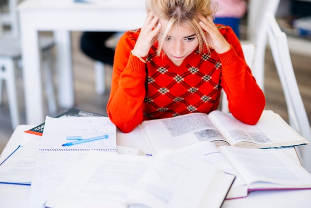A close-up shot of a stressed college student sitting at a desk piled high with books and papers, hands on their head, looking overwhelmed.