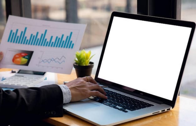 A person using a laptop, with charts and graphs displayed on the screen, in a brightly lit, minimalist home office. A cup of coffee and a notebook are also visible on the desk, conveying a sense of focused financial planning.